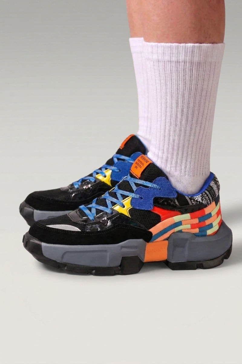 Baskets chunky 'Tsuruga' coloris noir — panneaux contrastés bleu et orange, semelle gomme, portées avec chaussettes blanches sur fond studio gris
