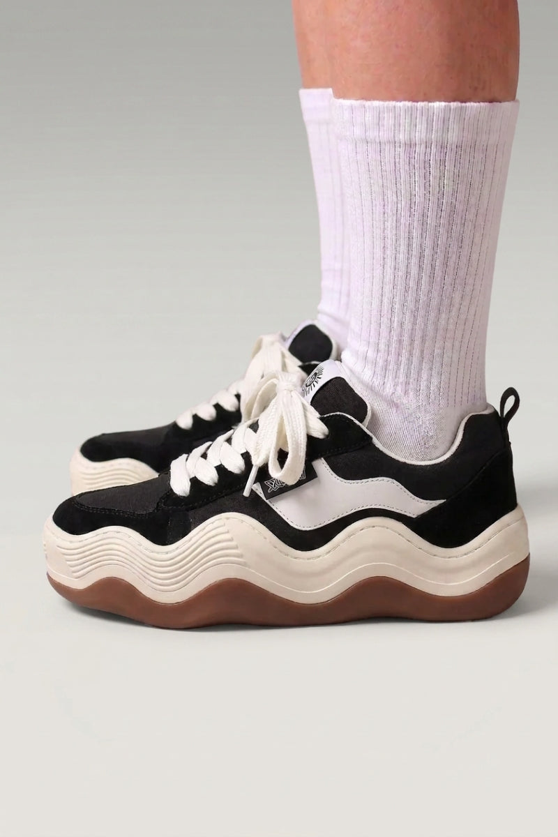 Baskets à plateforme 'Nami' coloris noir — semelle plateforme découpée en vague ondulée bicolore, portées avec chaussettes blanches sur fond studio gris