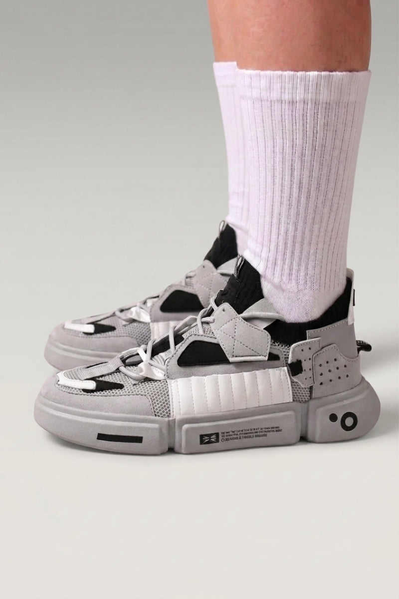 Baskets basses 'Kazuno' coloris gris — sangles utilitaires, œillets métalliques, semelle segmentée, portées avec chaussettes blanches sur fond studio gris