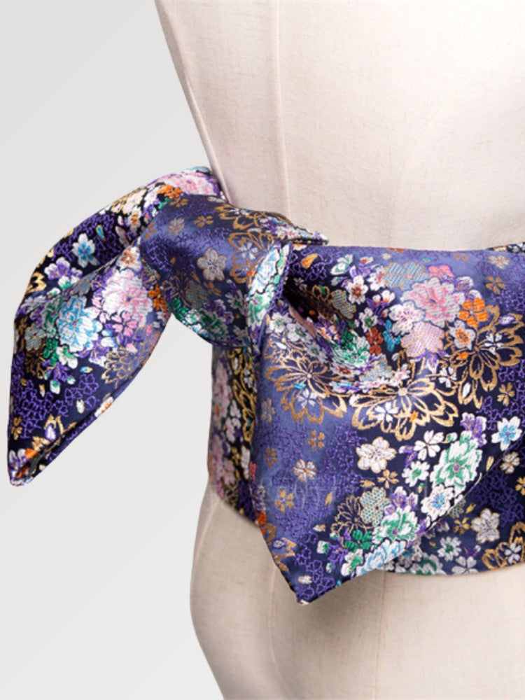 Gros plan du nœud de la ceinture obi 'Nadeshiko' sur un buste de mannequin — fond violet profond couvert de fleurs polychromes rehaussées d'or