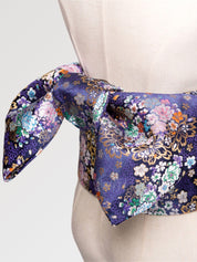 Gros plan du nœud de la ceinture obi 'Nadeshiko' sur un buste de mannequin — fond violet profond couvert de fleurs polychromes rehaussées d'or