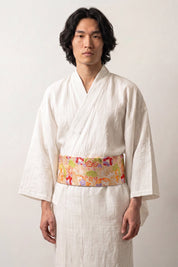 Ceinture obi 'Nanten' portée à la taille sur un kimono blanc par un mannequin homme — fond jaune d'or couvert de fleurs multicolores et de motifs éventail