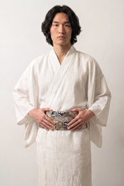 Ceinture obi 'Tachibana' portée à la taille sur un kimono blanc par un mannequin homme — brocart noir couvert de fleurs argentées et dorées en camaïeu