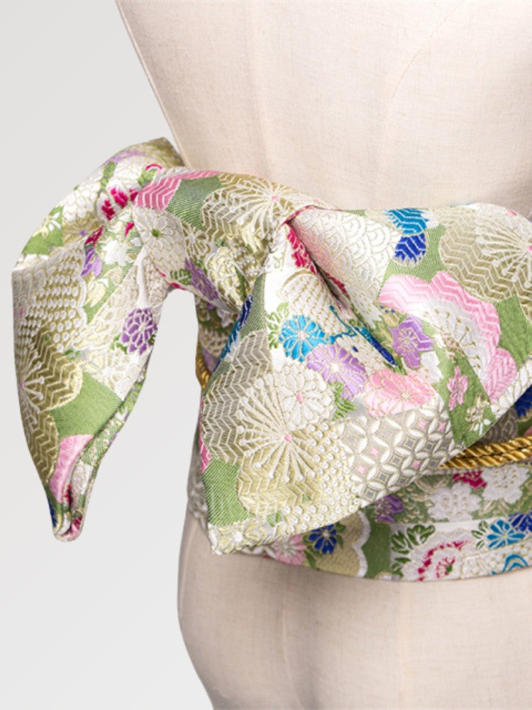 Gros plan du nœud de la ceinture obi 'Sakura' sur un buste de mannequin — brocart vert pâle et doré semé de fleurs roses et de grues