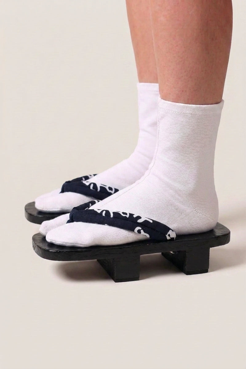 Geta japonaise 'Nagano' portée avec des chaussettes tabi blanches, vue de profil — plateforme en bois noirci, lanière bleu nuit à volutes blanches