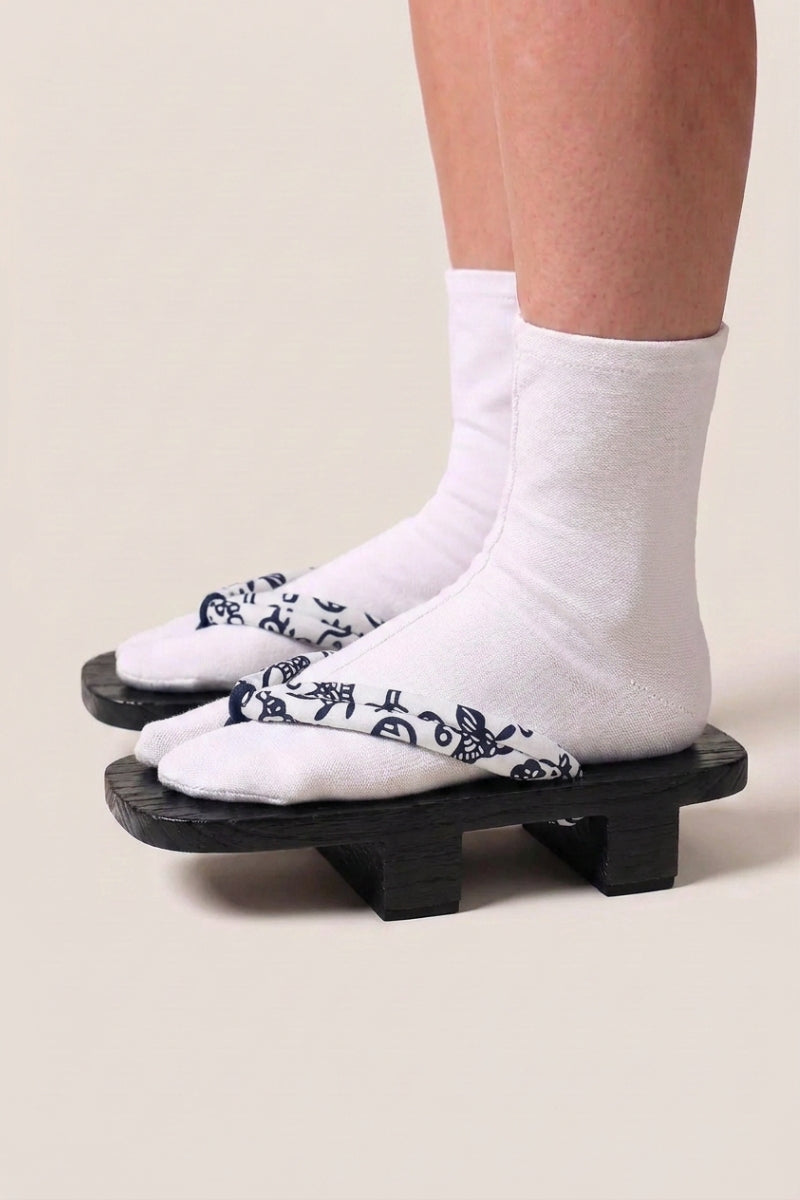 Geta japonaise 'Utsomita' portée avec des chaussettes tabi blanches, vue de profil — plateforme en bois noirci, lanière blanche à rinceaux bleu nuit