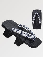 Paire de geta 'Utsomita' vue de trois quarts — socle en bois noirci à deux dents, lanière blanche à rinceaux bleu nuit, dessous visible