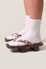 Geta japonaise 'Shakori' portée avec des chaussettes tabi blanches, vue de profil — plateforme en bois foncé, lanière rouge profond à motif cachemire blanc