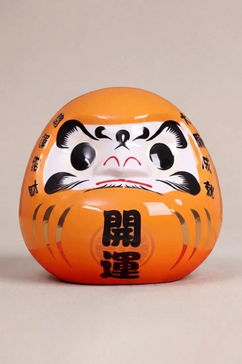 Daruma orange impérial en céramique posé sur fond beige — figurine ronde de 11 cm au kanji 皇, yeux vierges pour le rite du vœu