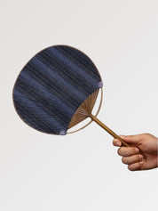 Éventail uchiwa japonais 'Shoji' modèle 1, tenu en main — toile tissée bleu nuit à fines rayures verticales, manche en bambou et cerclage rond
