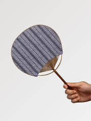 Éventail uchiwa japonais 'Shoji' modèle 2, tenu en main — toile tissée gris-bleu clair à rayures serrées, manche en bambou et cerclage rond
