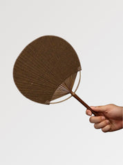 Éventail uchiwa japonais 'Shoji' modèle 3, tenu en main — toile tissée brun tabac unie au grain apparent, manche en bambou et cerclage rond