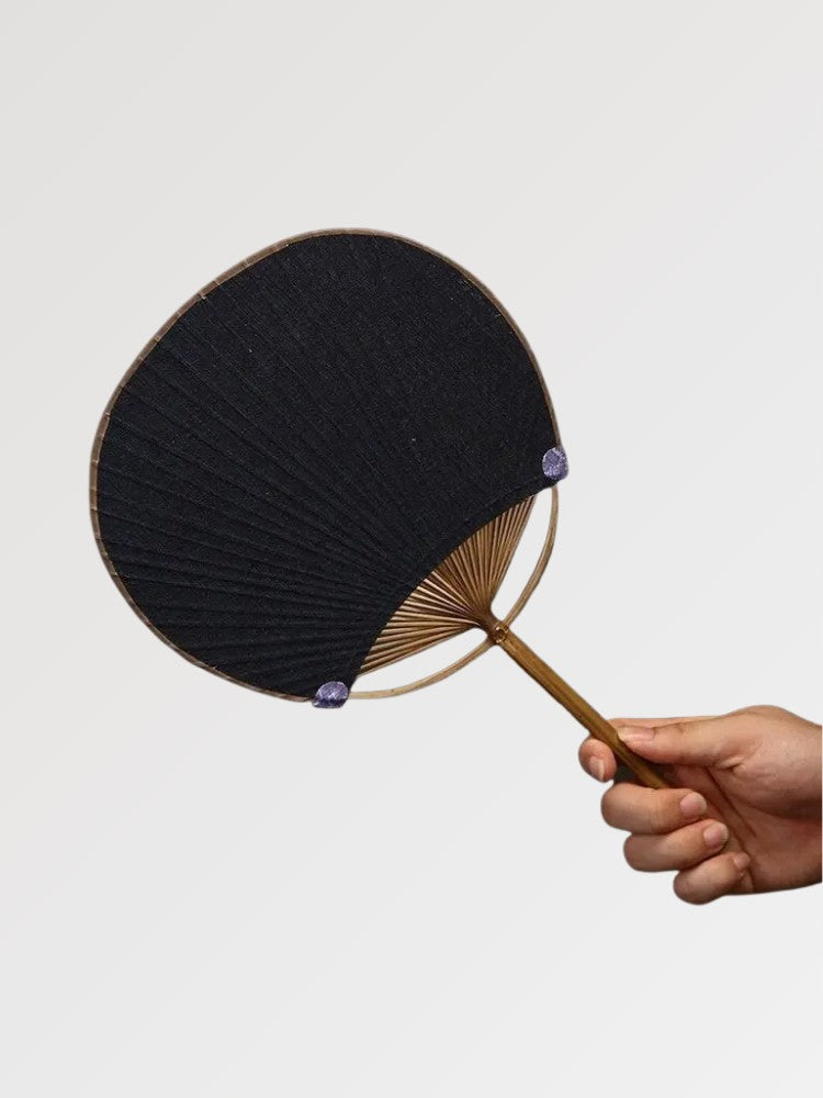 Éventail uchiwa japonais 'Shoji' modèle 4, tenu en main — toile tissée noire dense, sans motif apparent, manche en bambou et cerclage rond