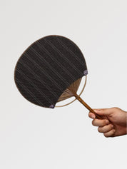 Éventail uchiwa japonais 'Shoji' modèle 5, tenu en main — toile tissée brun sombre à rayures verticales discrètes, manche en bambou et cerclage rond