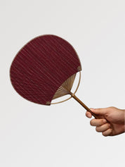 Éventail uchiwa japonais 'Shoji' modèle 8, tenu en main — toile tissée bordeaux profond au grain marqué, manche en bambou et cerclage rond