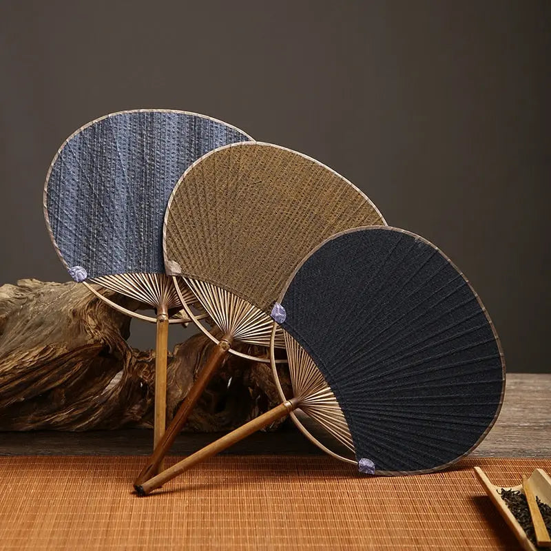 Trois éventails uchiwa 'Shoji' posés contre un bois flotté — toiles tissées bleu, brun et beige, cerclages ronds en bambou