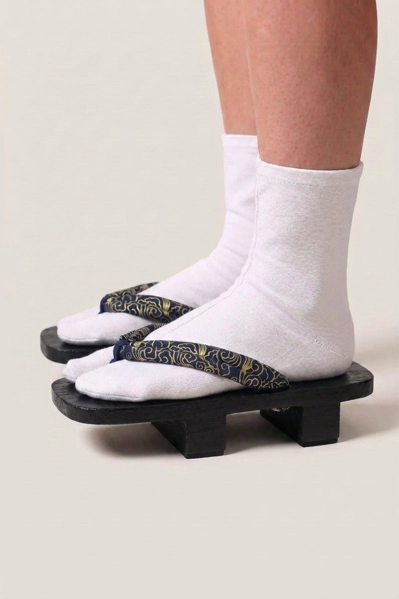 Geta japonaise 'Tokoname' portée avec des chaussettes tabi blanches, vue de profil — plateforme en bois noirci, lanière sombre à arabesques dorées