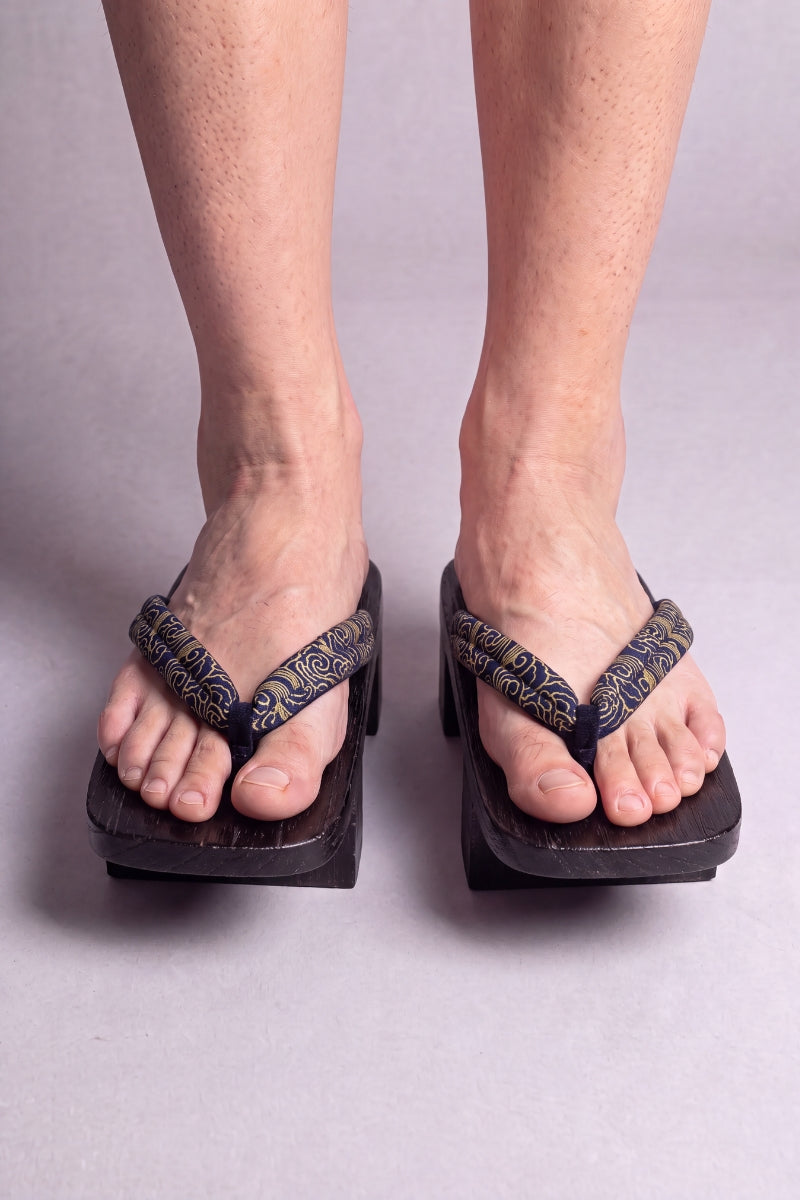 Geta japonaise 'Tokoname' portée pieds nus, vue de face — lanière sombre à arabesques dorées sur plateforme surélevée en bois noirci