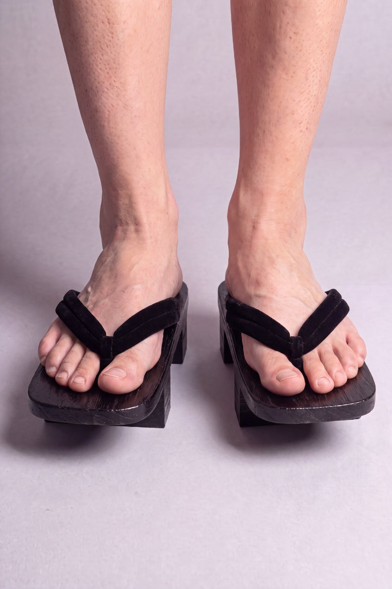 Geta japonaise 'Hekinan' portée pieds nus, vue de face — lanière noire mate assortie au socle sur plateforme surélevée en bois noirci
