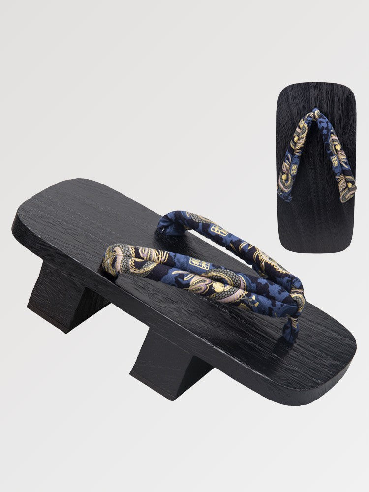 Paire de geta 'Nagoya' vue de trois quarts — socle en bois noirci à deux dents, lanière bleu nuit à rinceaux dorés, dessous visible