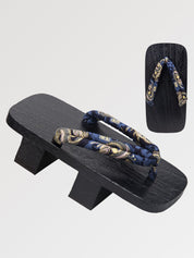 Paire de geta 'Nagoya' vue de trois quarts — socle en bois noirci à deux dents, lanière bleu nuit à rinceaux dorés, dessous visible