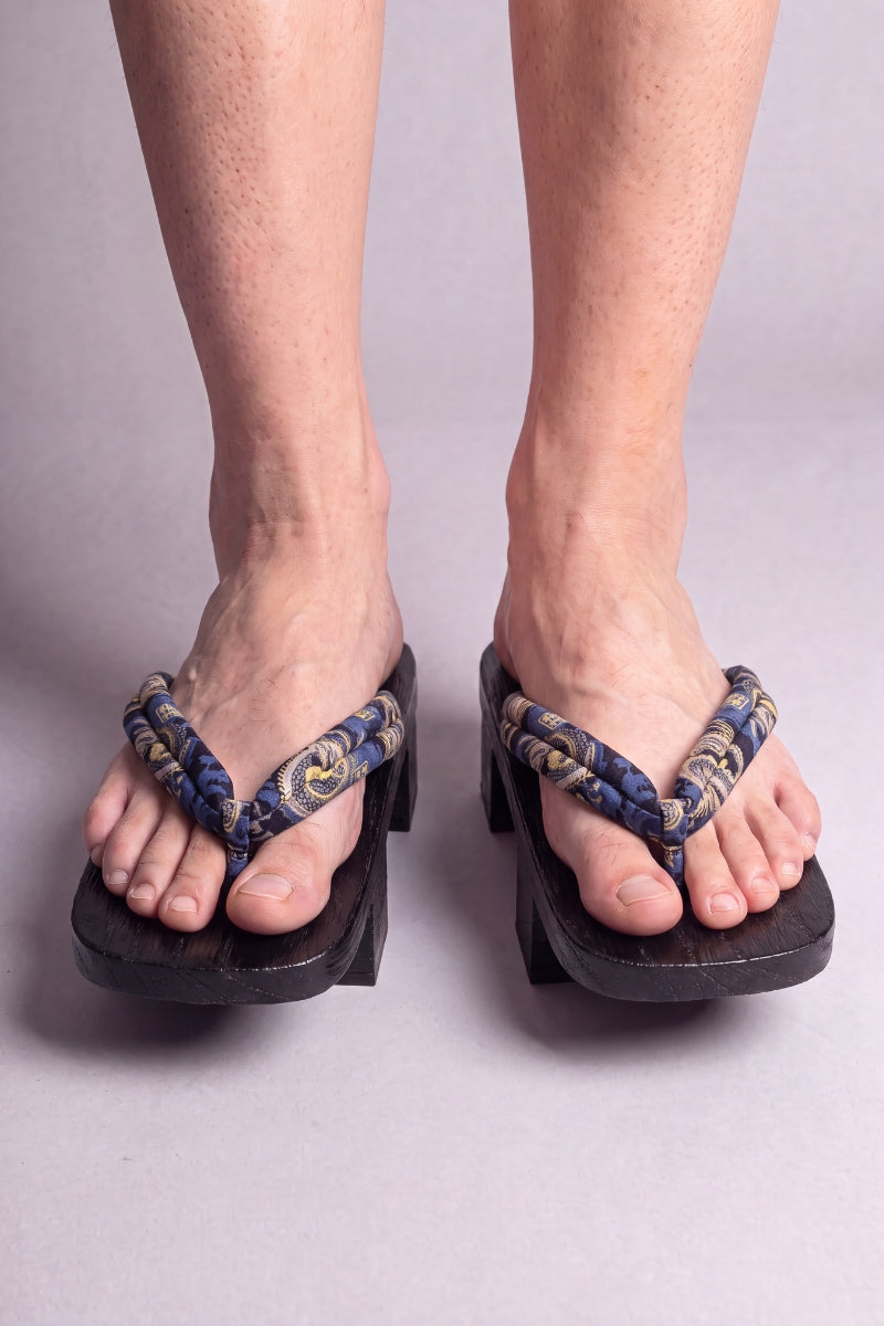 Geta japonaise 'Nagoya' portée pieds nus, vue de face — lanière bleu nuit à rinceaux dorés sur plateforme surélevée en bois noirci