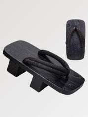 Paire de geta 'Tsushima' vue de trois quarts — socle en bois noirci à deux dents, lanière noire fine et souple, dessous visible