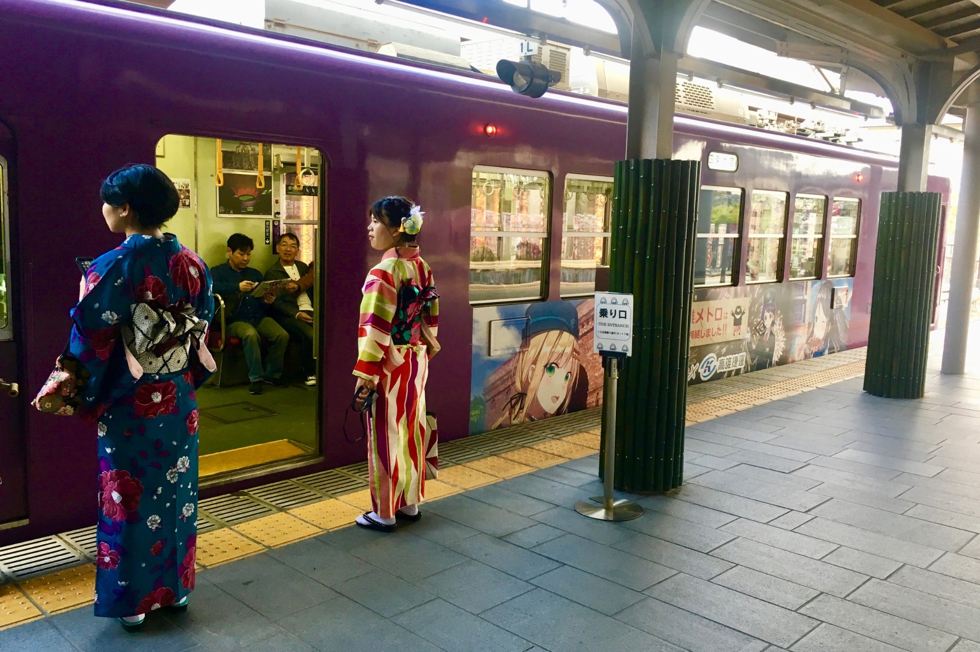 Deux femmes en kimono attendant un train violet décoré d’illustrations d’anime à Kyoto, entre tradition japonaise et culture contemporaine.