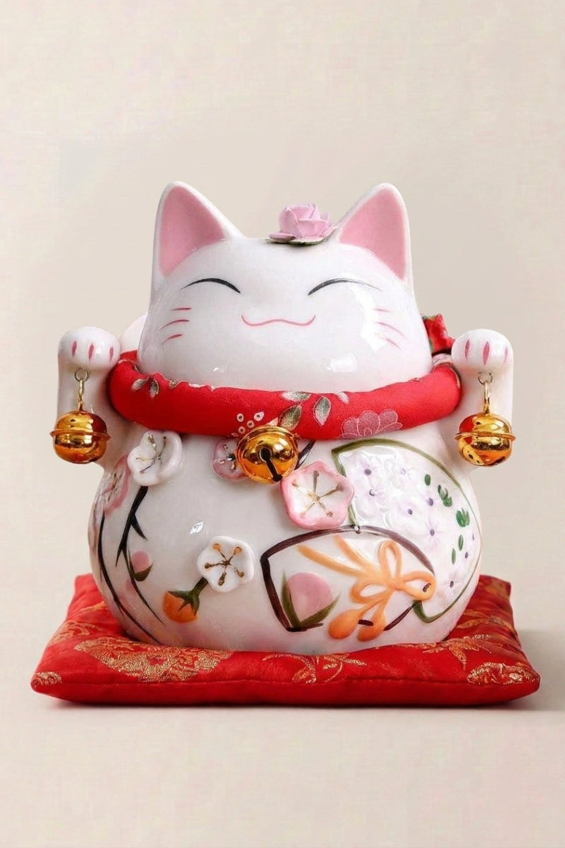 Maneki neko tirelire 'Okami' en céramique — chat blanc aux yeux fermés, ventre orné de motifs floraux et grelots dorés, sur un coussin rouge
