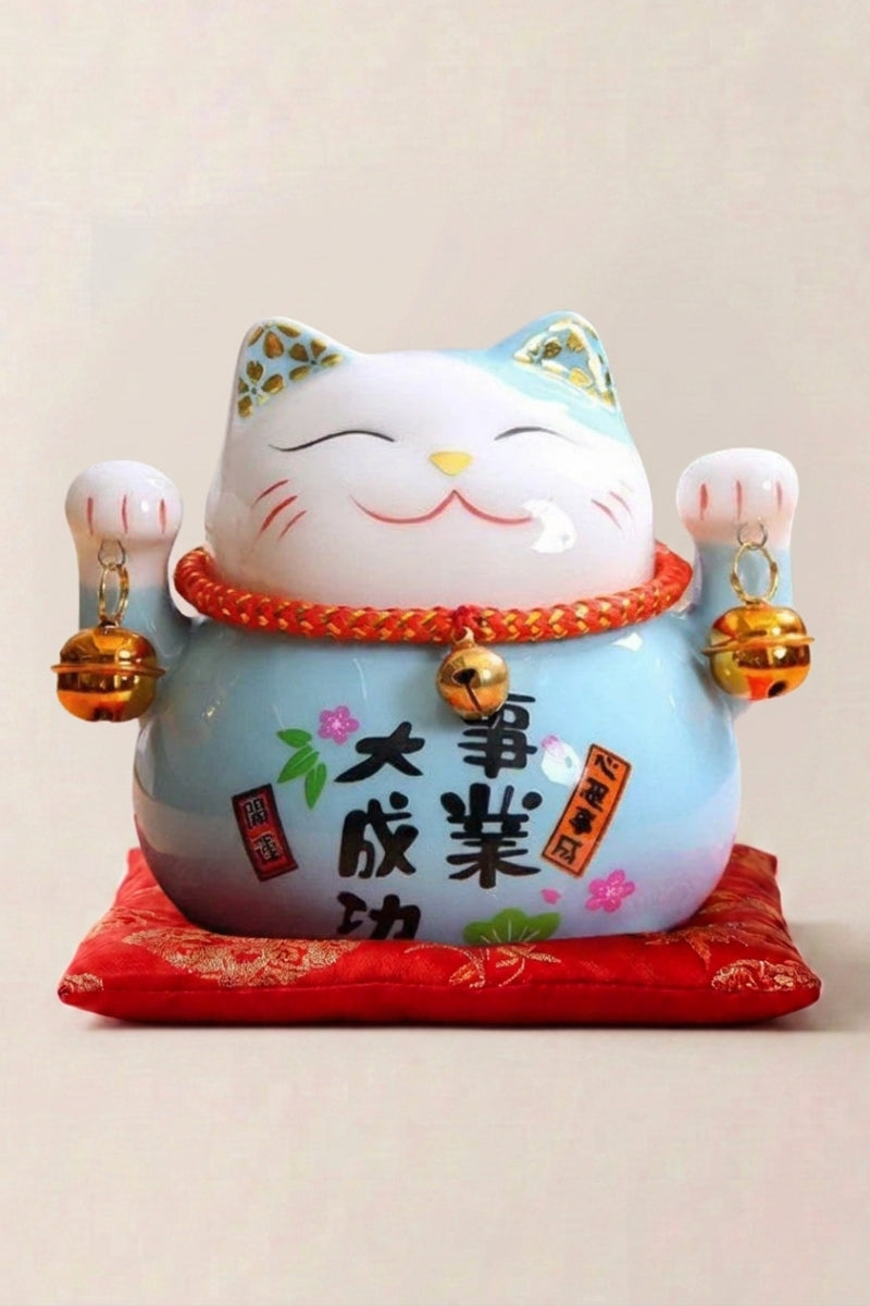 Maneki neko tirelire 'Blue' en céramique — chat bleu ciel à plaque calligraphiée et grelot doré, les deux pattes levées, sur un coussin rouge