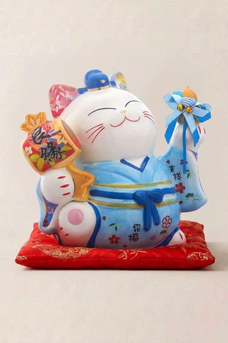 Maneki neko tirelire 'Blue Fortune' en céramique — chat bleu en kimono à nœud, sac de fortune sur l'épaule et patte droite levée, sur un coussin rouge