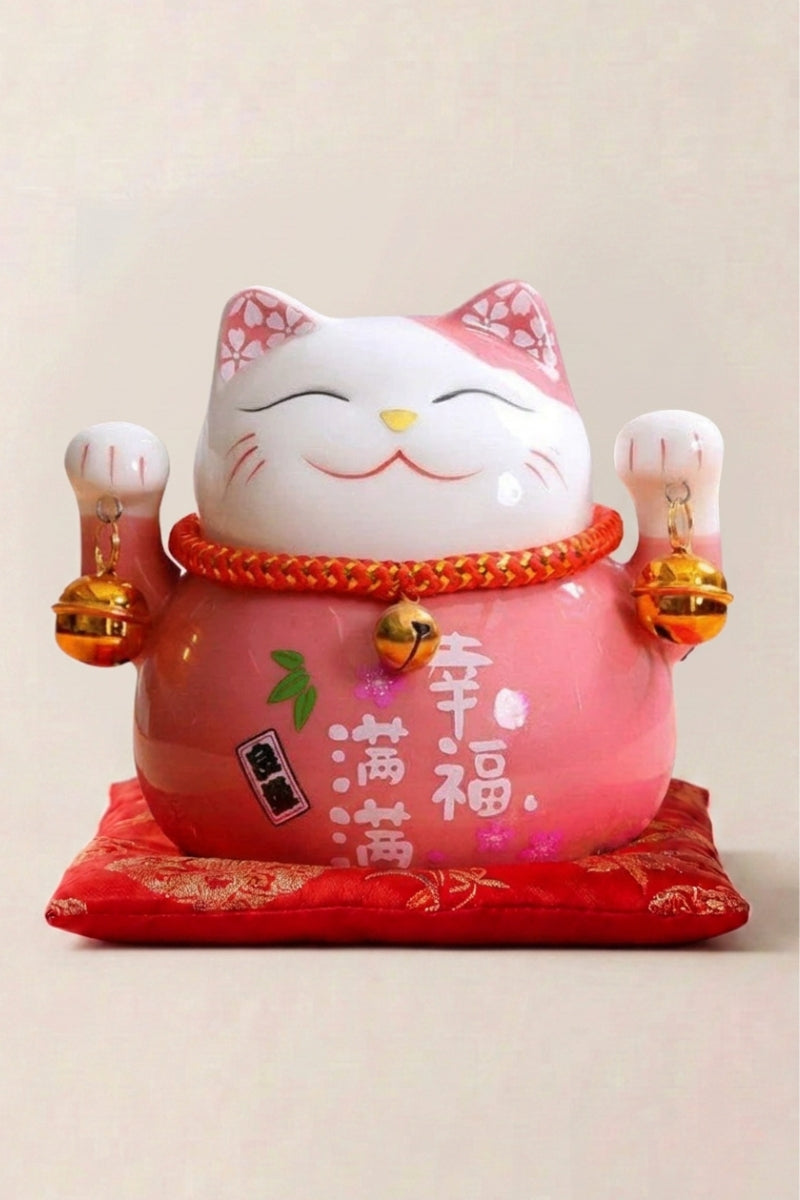Maneki neko tirelire 'Rose' en céramique — chat rose pâle à fleurs de cerisier et plaque calligraphiée, les deux pattes levées, sur un coussin rouge