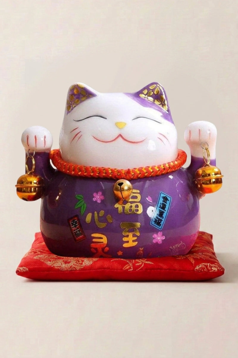 Maneki neko tirelire 'Violet' en céramique — chat violet à décor doré, les deux pattes levées, assis sur un coussin rouge, sur un coussin rouge