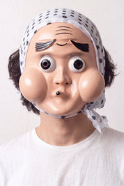 Masque hyottoko porté par un mannequin sur fond clair — visage comique aux joues rondes, bouche pincée de travers et bandeau bleu