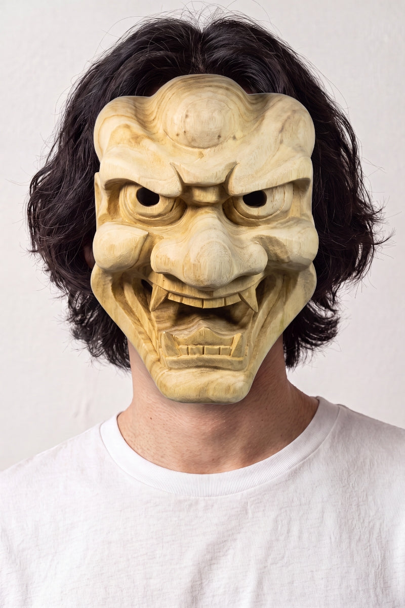 Masque oni en bois sculpté porté par un mannequin sur fond clair — démon aux sourcils broussailleux et aux crocs saillants, bois clair veiné