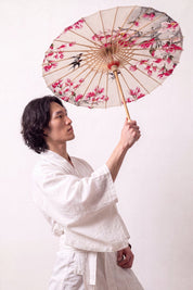 Ombrelle japonaise 'Nomikomu' ouverte et tenue par un mannequin en kimono blanc — toile crème couverte de fleurs de cerisier roses