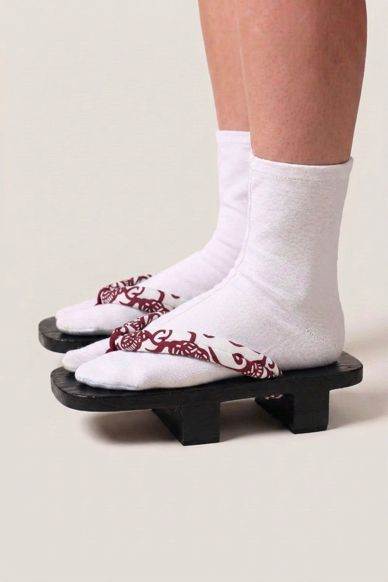 Geta japonaise 'Aomori' portée avec des chaussettes tabi blanches, vue de profil — plateforme en bois noirci, lanière rouge et blanche à motif cachemire