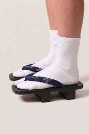 Geta japonaise 'Okimari' portée avec des chaussettes tabi blanches, vue de profil — plateforme en bois noirci, lanière bleu nuit à motif floral blanc