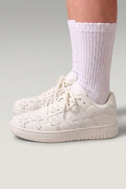 Baskets basses 'Shiro' coloris blanc — croix embossées sur les flancs, lacets épais assortis, portées avec chaussettes blanches sur fond studio gris