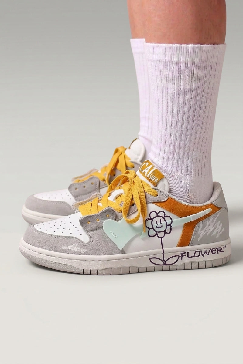 Baskets basses 'Hanabi' coloris blanc — patchs colorés, fleur cousue au talon, lacets larges, portées avec chaussettes blanches sur fond studio gris