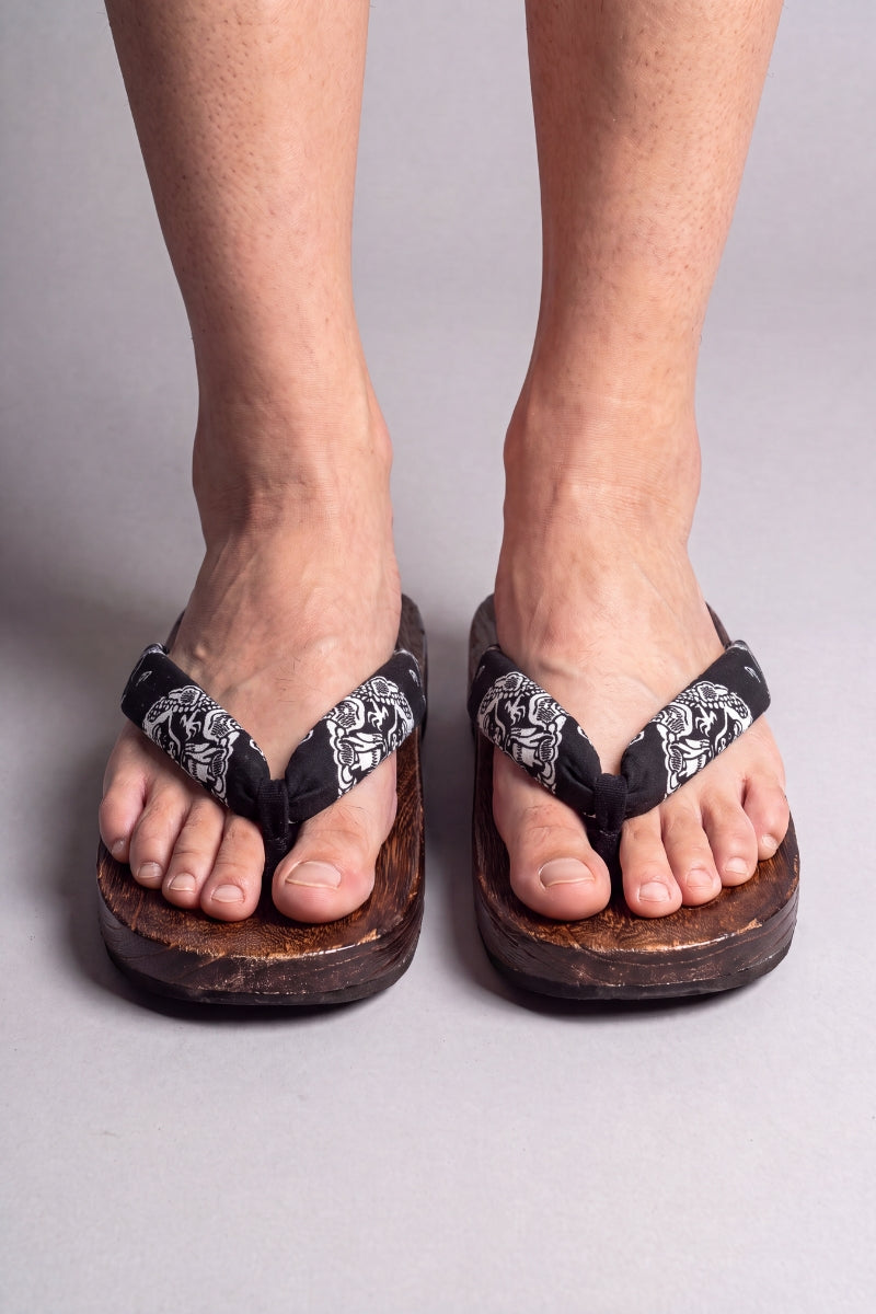 Tongs japonaises coloris fleurs blanches sur noir, portées pieds nus de face — semelle en bois massif brun et lanière rembourrée passant entre les orteils