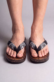 Tongs japonaises coloris fleurs blanches sur noir, portées pieds nus de face — semelle en bois massif brun et lanière rembourrée passant entre les orteils