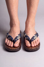 Tongs japonaises coloris fleurs dorées sur bleu nuit, portées pieds nus de face — semelle en bois massif brun et lanière rembourrée passant entre les orteils