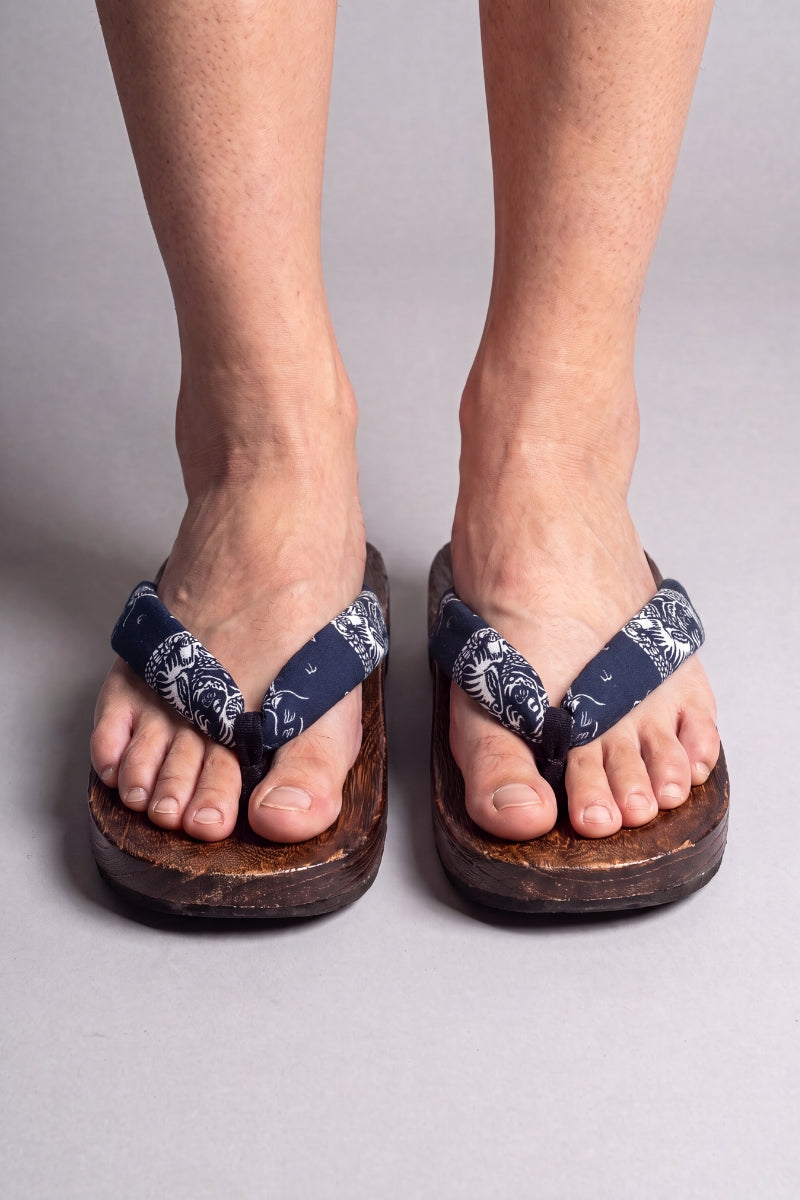 Tongs japonaises coloris fleurs blanches sur bleu nuit, portées pieds nus de face — semelle en bois massif brun et lanière rembourrée passant entre les orteils