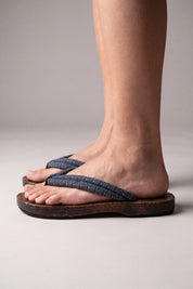 Tongs japonaises vues de profil, portées pieds nus — semelle en bois plate et galbée, lanière bleu-gris à motif de vagues